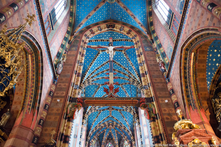 Upward view of blue star-patterned Gothic vaulting, painted ribs and walls, clerestory windows, and a large suspended crucifix.