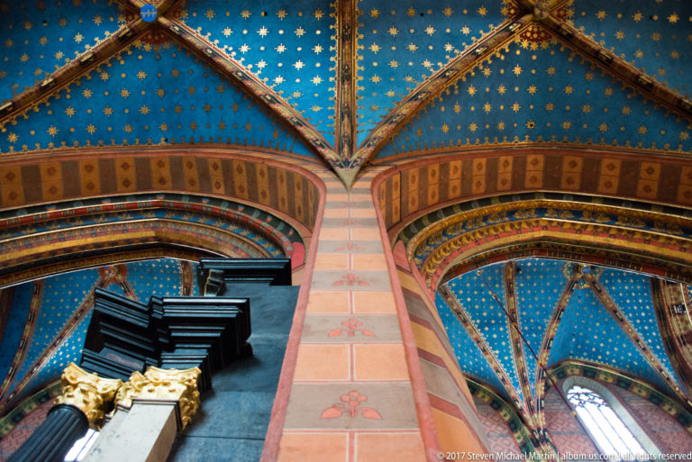 Close upward view of blue starry vault cells and painted ribs above a tall pier, with a dark organ case at lower left.