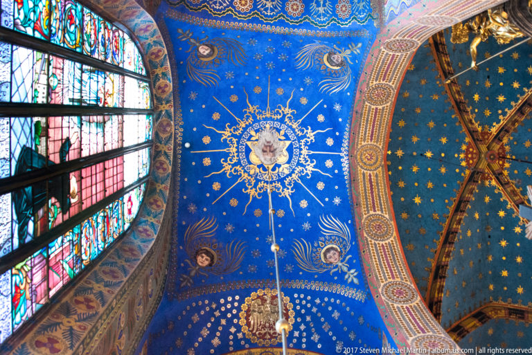 Circular sacred medallion surrounded by gold ornament and stars on a blue vault, bordered by stained glass and a painted arch.