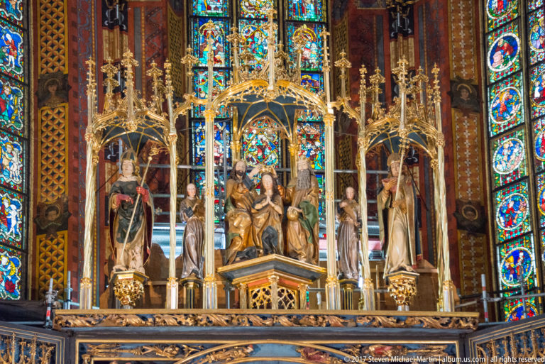 Gilded shrine with a seated central figure and standing attendants beneath Gothic canopies, framed by bright stained-glass windows.