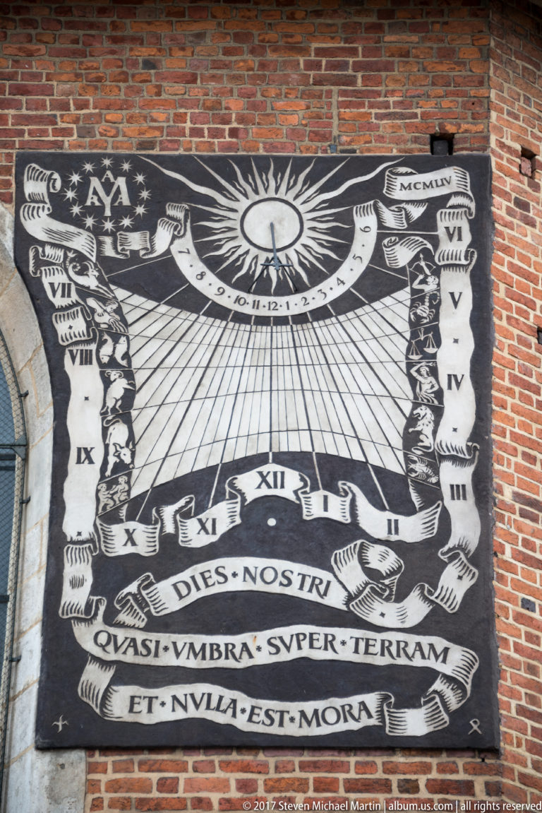 Large black-and-white sundial with Roman numerals, radiating hour lines, solar emblem, and Latin text mounted on a red-brick church wall.