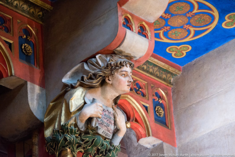 Polychrome angel with flowing hair holding an inscribed tablet or book, set against blue, red, and gold painted architectural decoration.