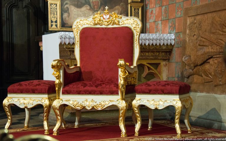 Red-upholstered gilded ceremonial chair on a red carpet, flanked by two matching low stools with carved gold frames.