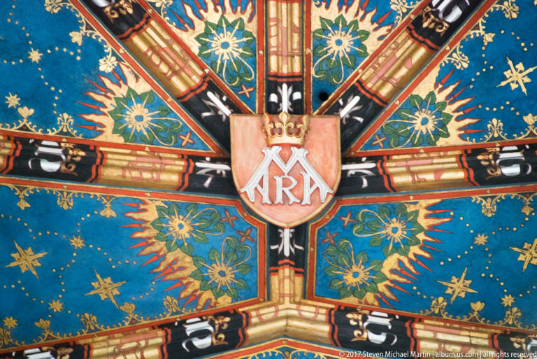 Shield-shaped monogram at the crossing of painted vault ribs, surrounded by flowers, stars, and ornament on a vivid blue ceiling.