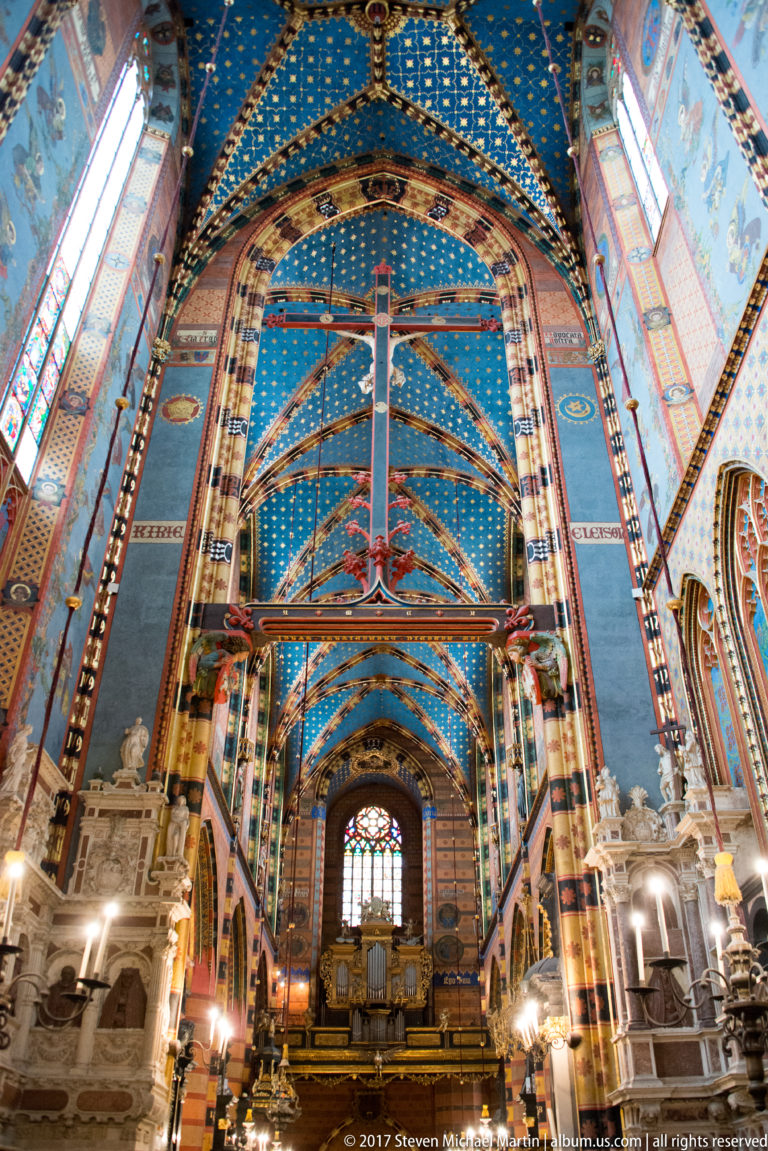 Axial nave view with a large suspended crucifix beneath blue starry vaulting, flanked by painted piers and side monuments.