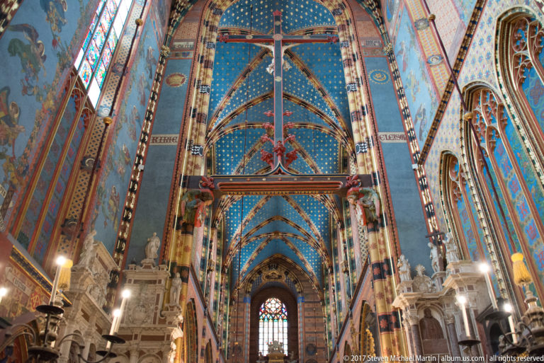 Steep upward nave view with suspended crucifix, blue star-filled vault bays, painted piers, and pale side monuments.