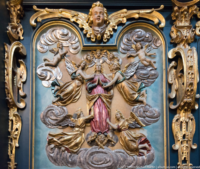 Polychrome relief of a crowned female sacred figure surrounded by angels and clouds within an elaborate gilded frame.