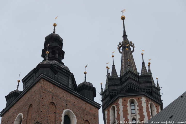 Upward view of St. Mary’s Basilica’s unequal brick towers with dark roofs, pinnacles, and gilded finials against a gray sky.