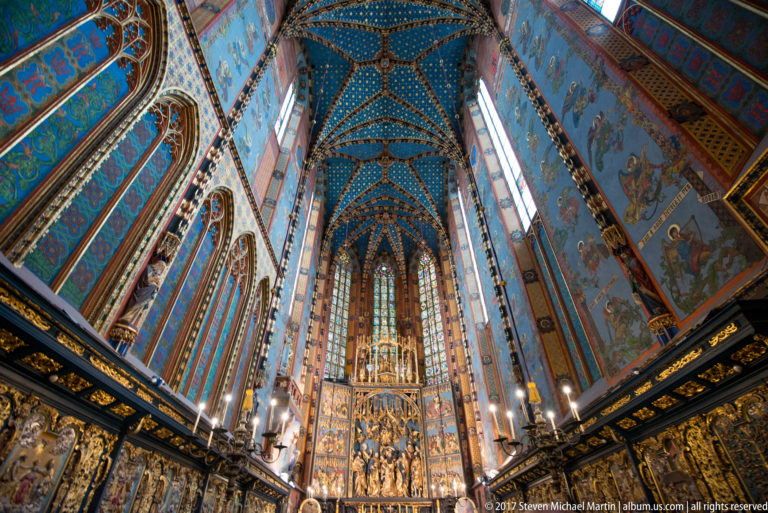 Wide vertical chancel view with the Veit Stoss Altarpiece, stained-glass windows, blue rib vaulting, and dark choir furnishings.
