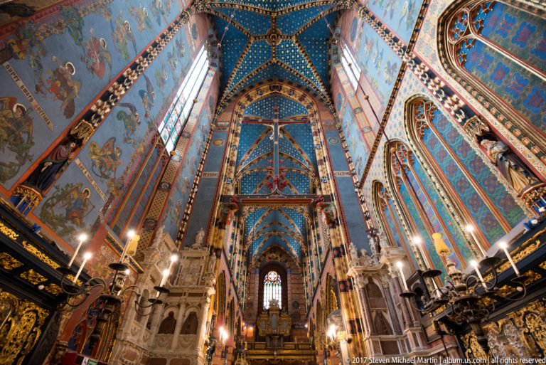 Long interior view of the nave with blue starry vaulting, striped piers, chandeliers, dark furnishings, and a distant gallery.