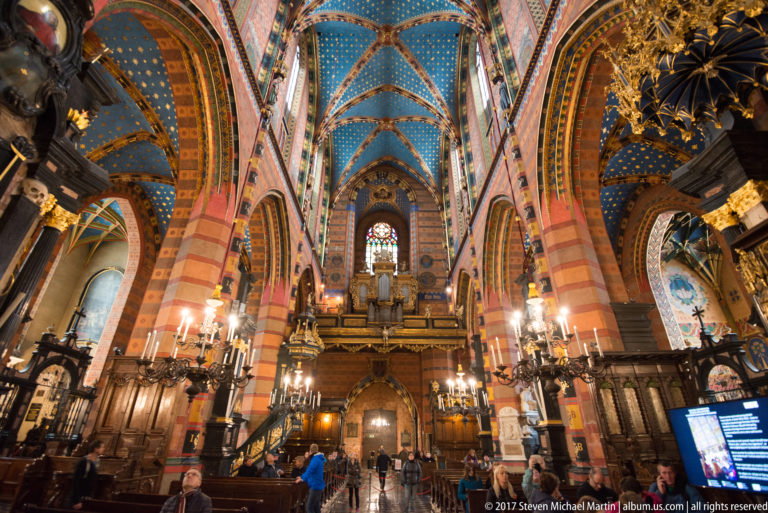 Broad nave view toward a raised gallery or organ loft, with striped piers, blue starry vaulting, chandeliers, and visitors below.