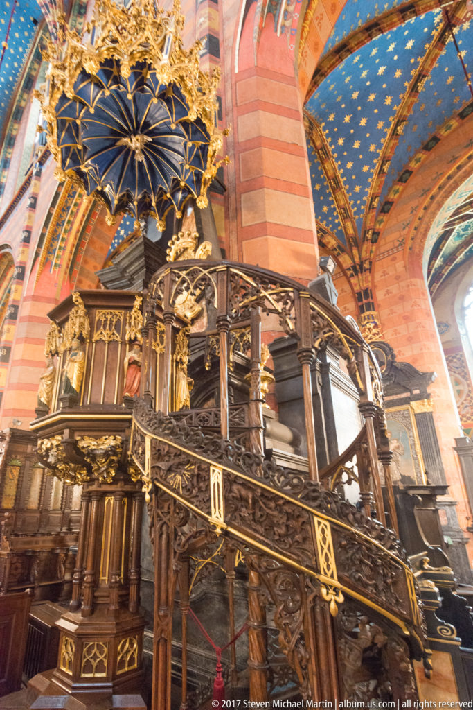 Ornate dark wooden pulpit with staircase, pierced railings, gilded details, and canopy beside a striped pier under blue vaulting.