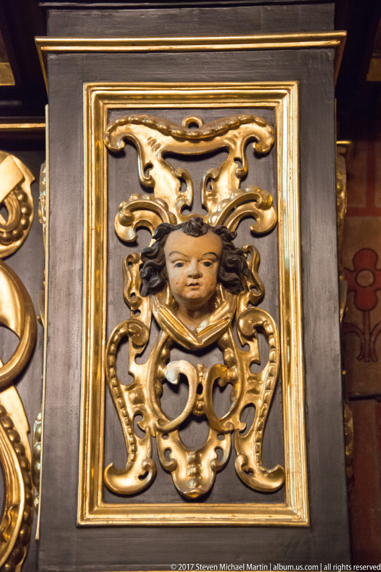 Close view of a gilded cherubic head within a scrolling cartouche mounted on a dark wooden panel.