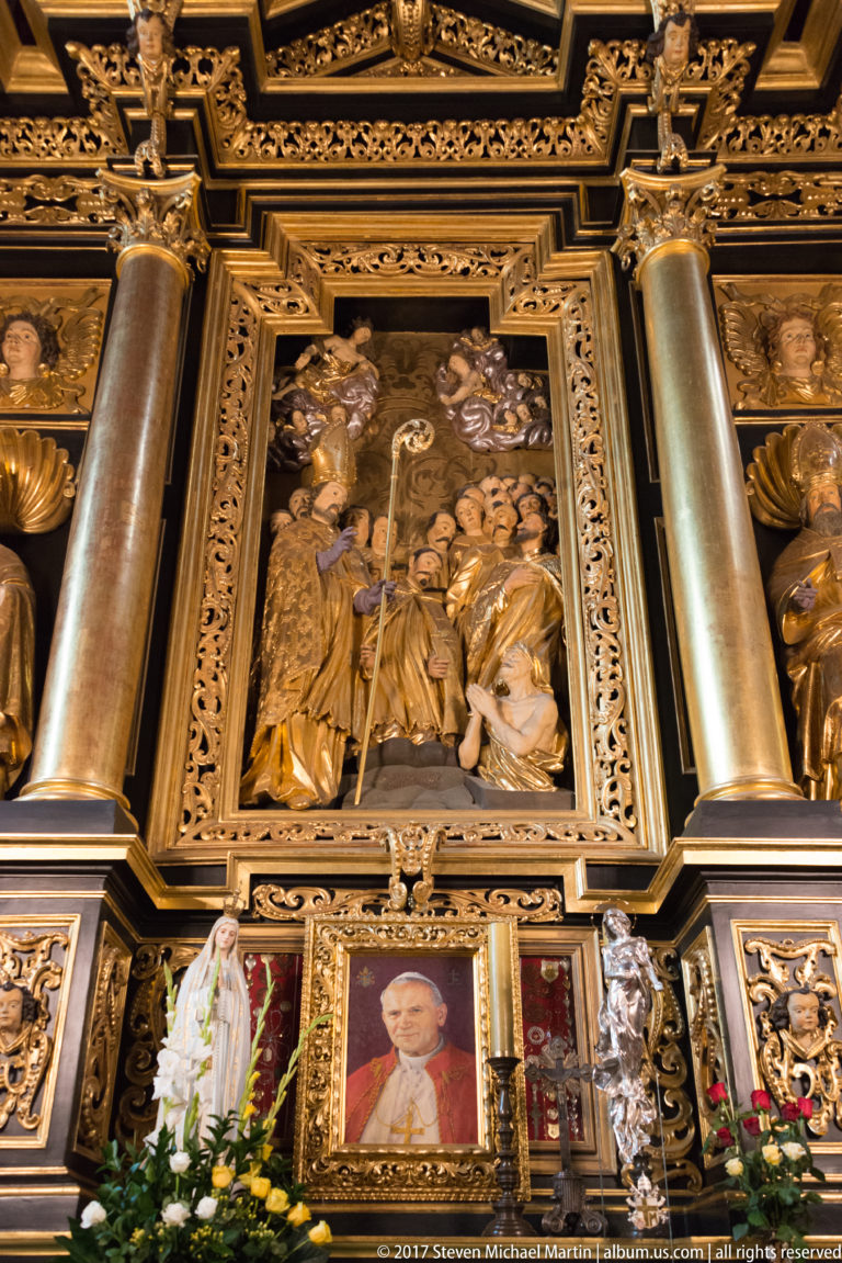 Dark-and-gold devotional altar with a sculpted relief scene above framed portraits, flowers, columns, and carved ornament.