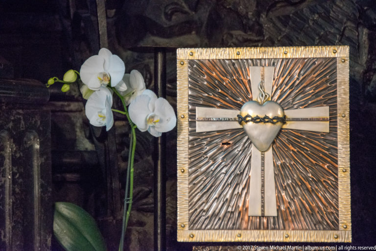 Radiant Sacred Heart emblem on a rectangular metal panel beside a white orchid, with rays spreading from the central heart.