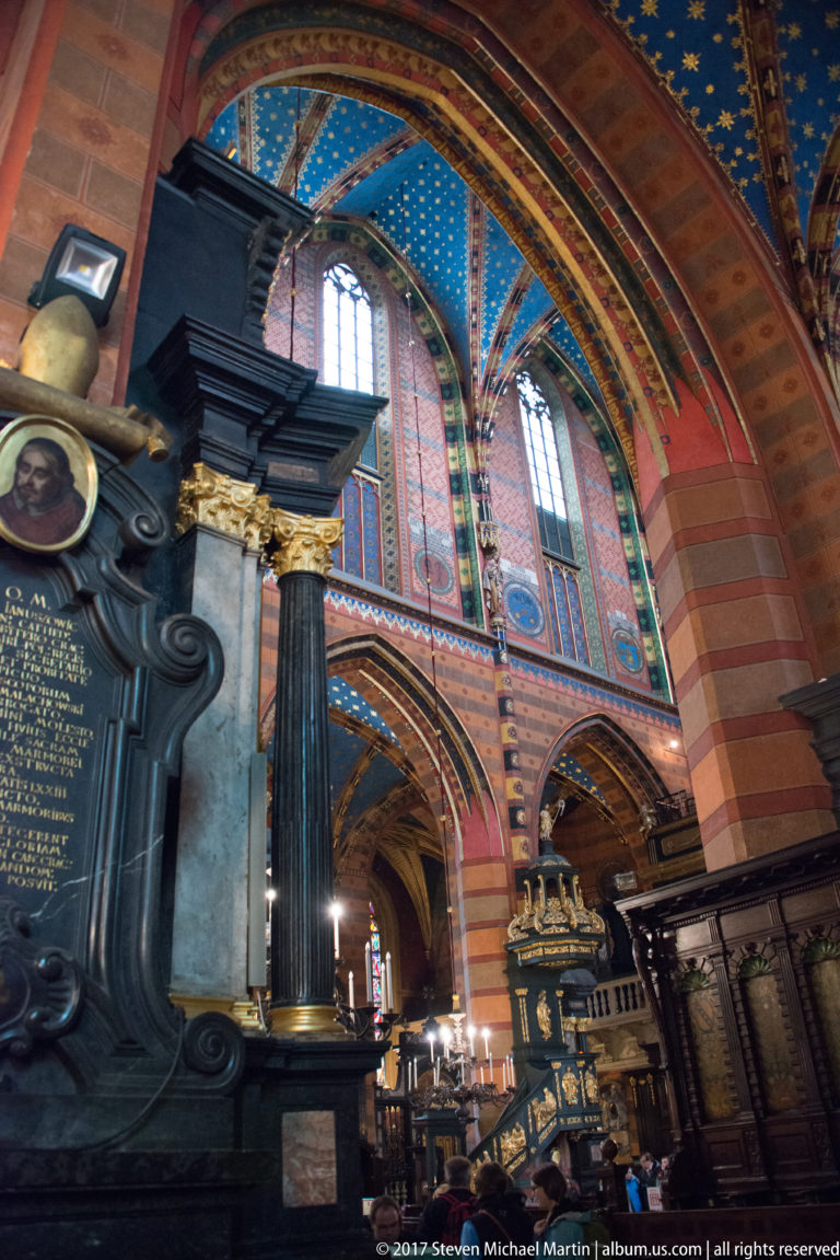 Dark memorial monument with oval portrait and gold inscription beside a black column in a richly painted Gothic side aisle.