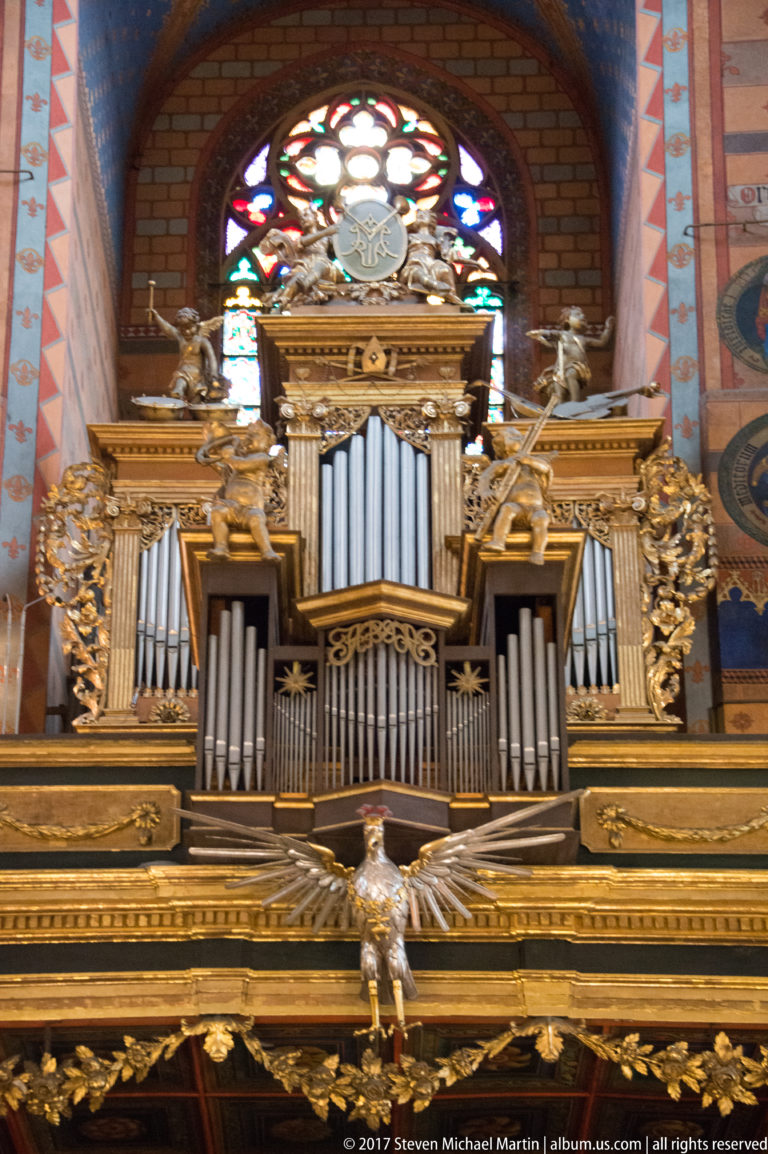 Ornate pipe organ with silver-toned pipes and gilded carved case beneath a stained-glass rose window.