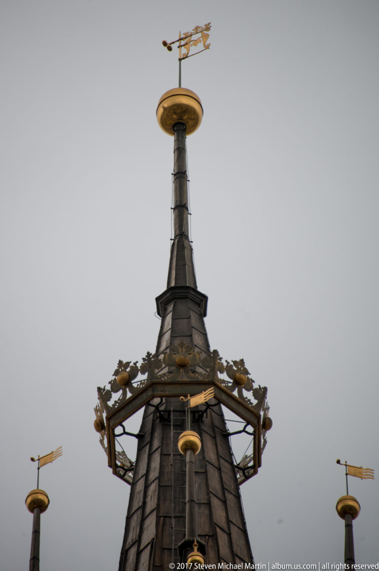 Close view of a dark metal church spire with gilded disk, weather vane, ornamental metalwork, and small projecting finials.