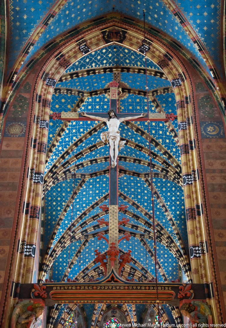 Large suspended crucifix beneath blue star-patterned vaulting and repeating Gothic arches inside St. Mary’s Basilica.