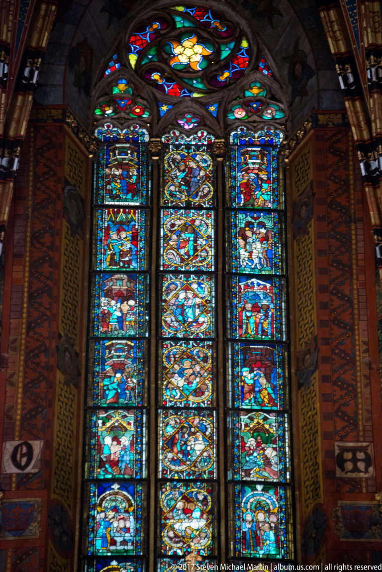 Three tall narrative stained-glass lancets beneath a circular window, framed by richly painted interior walls.