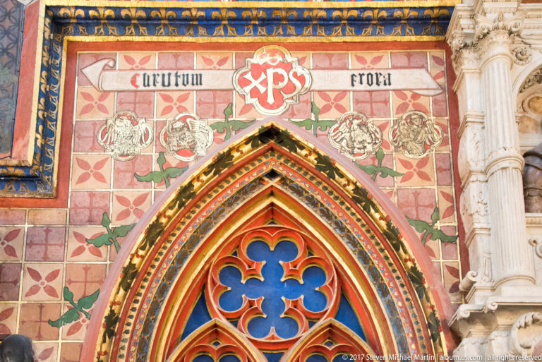 Polychrome pointed arch with geometric ornament, a Christogram-like monogram, Latin inscription, and floral motifs above.