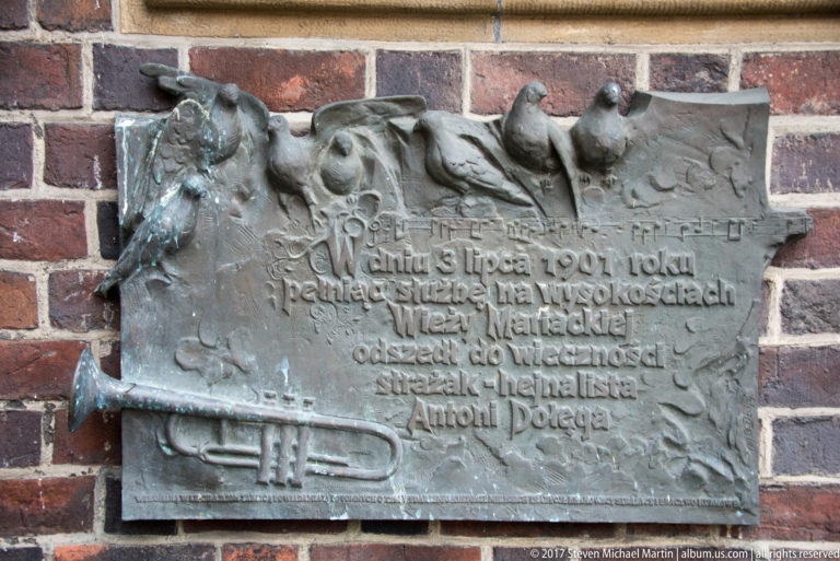 Weathered bronze memorial plaque with raised Polish text and relief forms mounted on the basilica’s red-brick exterior.