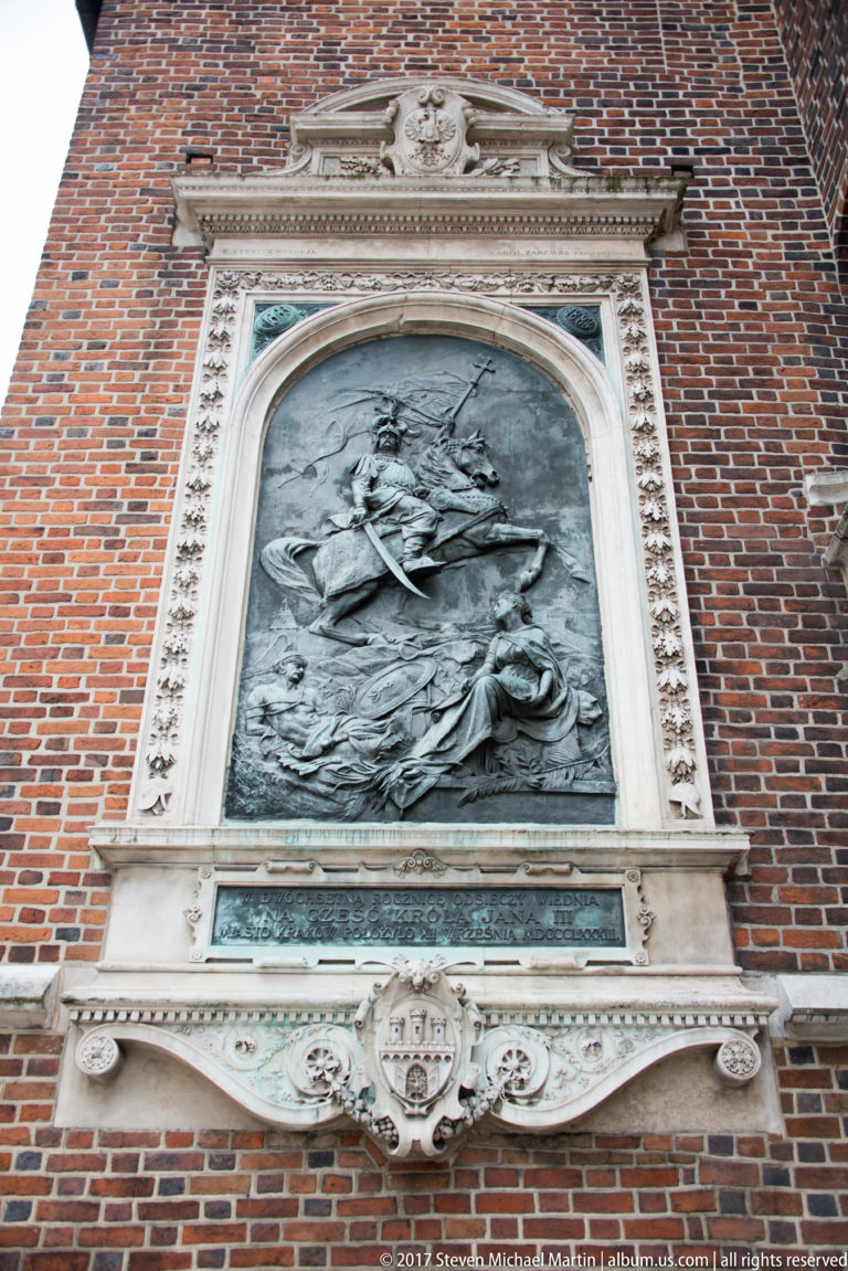 Large bronze multi-figure relief in a pale stone frame mounted high on a red-brick church wall, with inscription plaque below.