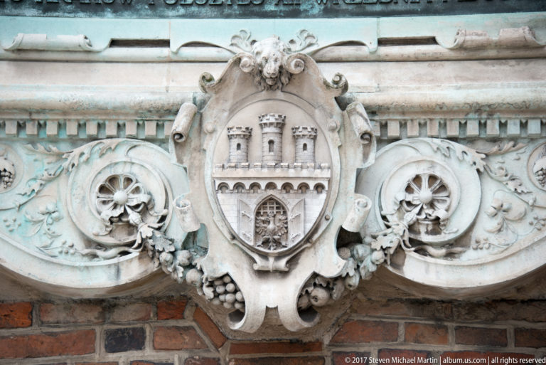 Carved stone cartouche with a three-towered gate shield, scrolling foliage, and rosettes mounted on a red-brick wall.