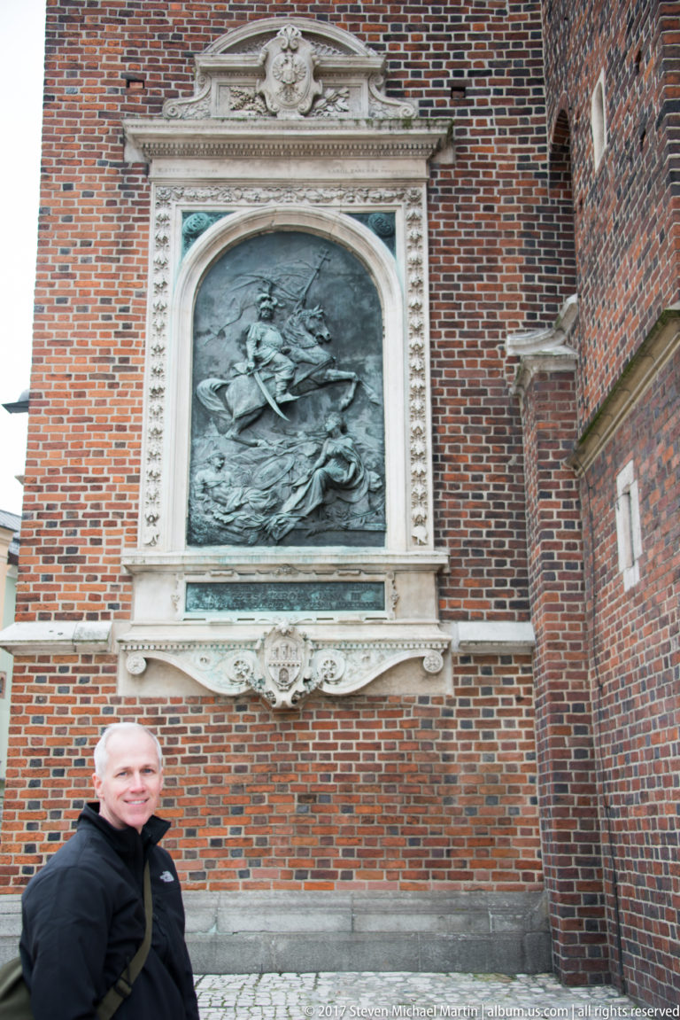 Visitor standing beneath a large bronze relief monument in a pale stone frame on the red-brick exterior of St. Mary’s Basilica.