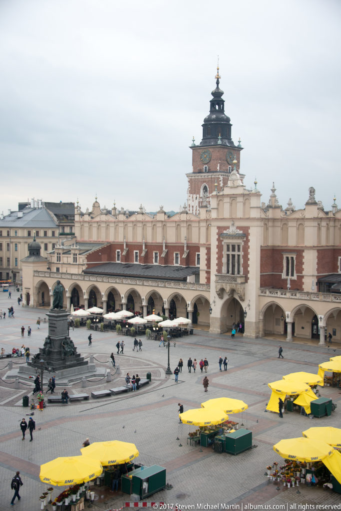 Elevated view across Krakow’s Main Market Square toward the arcaded Cloth Hall and tall Town Hall Tower, with pedestrians and yellow umbrellas.