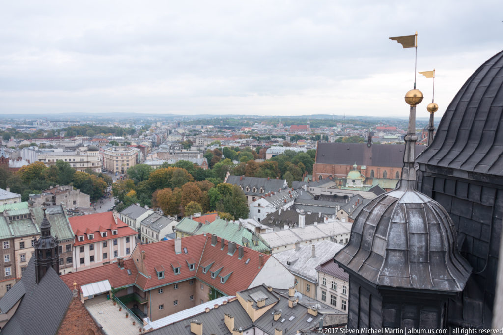 Dark metal basilica roof forms and gilded finials in the foreground above a broad panorama of Krakow’s rooftops.