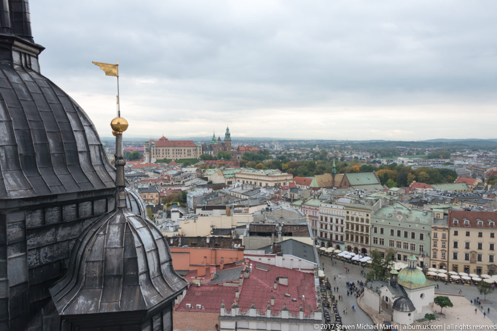 Elevated Krakow panorama framed by a dark metal basilica roof and gilded finial, with historic towers and rooftops beyond.