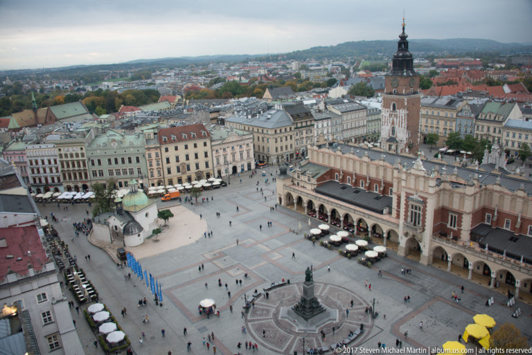 Wide elevated panorama of Rynek Główny with Cloth Hall, Town Hall Tower, historic façades, café seating, and pedestrians.