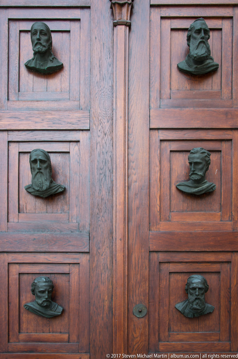 Paneled wooden church doors decorated with multiple projecting dark-metal portrait heads, each set within a separate rectangular panel.