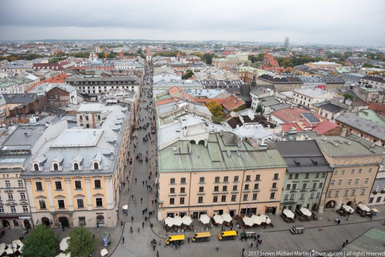 Elevated view along a busy straight Krakow Old Town street lined with historic townhouses, pedestrians, café seating, and mixed rooftops.