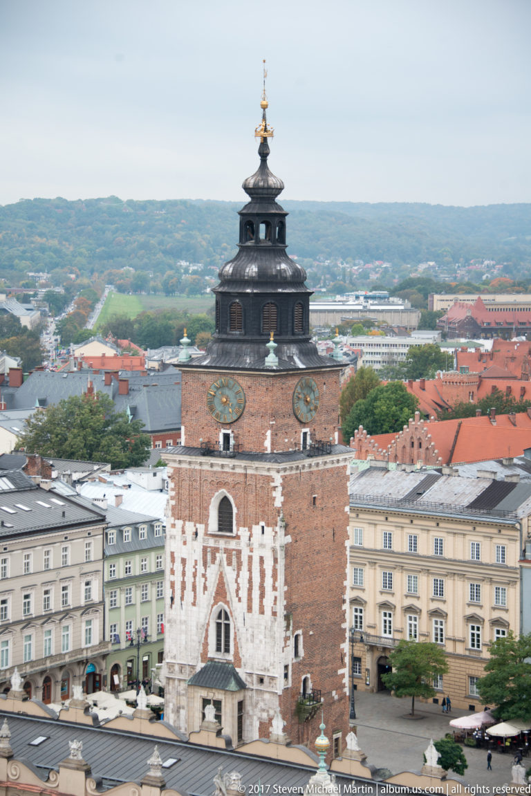 Elevated view of Krakow Town Hall Tower with clock face, dark tiered roof, gilded finial, and historic square façades below.