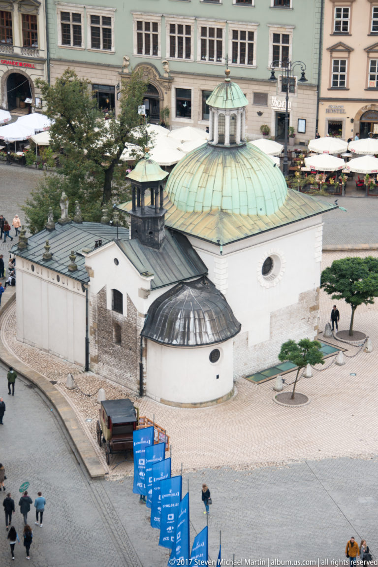 Elevated view of the small white Church of St. Adalbert with green-patinated dome, surrounded by pedestrians on Main Market Square.