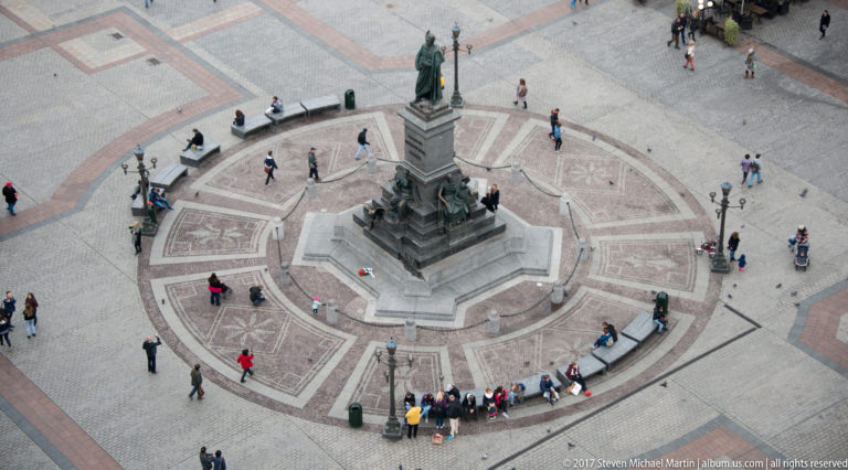 Elevated view of the Adam Mickiewicz Monument on a circular paved setting, with visitors gathered around the stone pedestal and bronze figures.