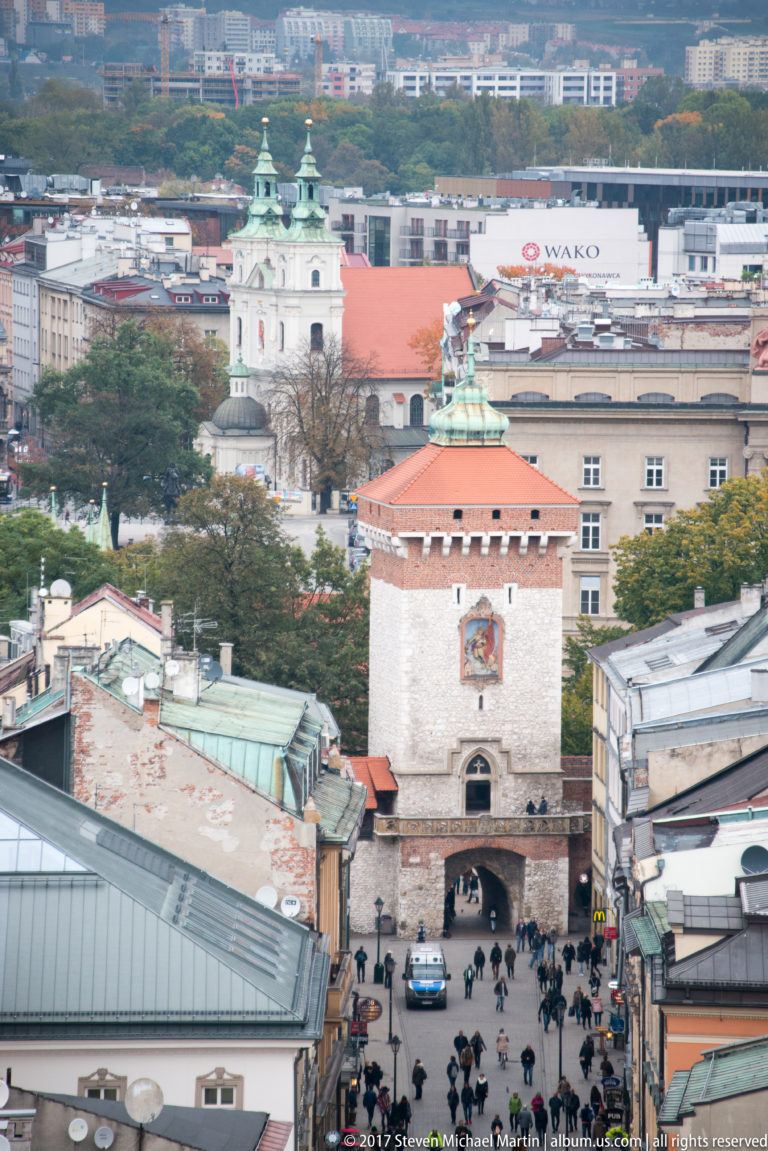 Elevated view along a busy Old Town street toward St. Florian’s Gate, with its red roof and arched passage framed by city buildings.