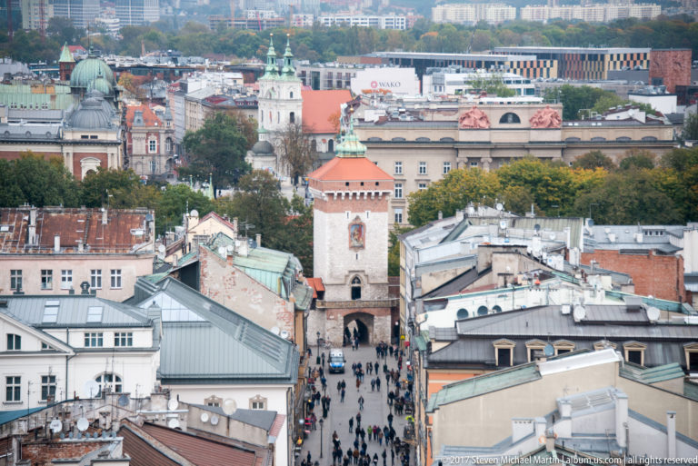 Long elevated view toward red-roofed St. Florian’s Gate, surrounded by dense historic rooftops and a distant city skyline.