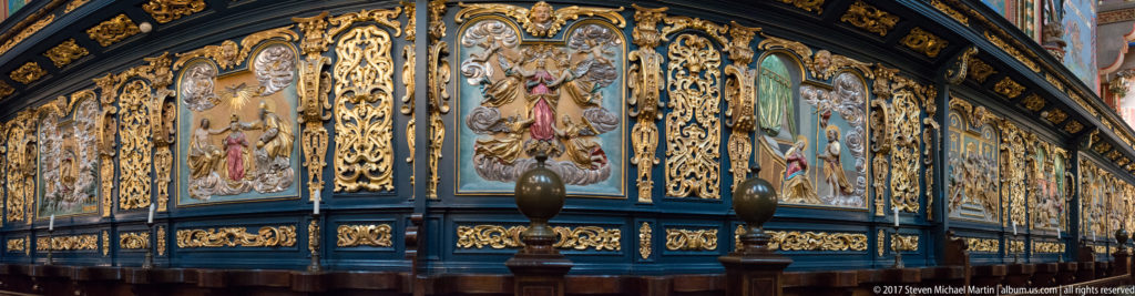 Oblique view of a dark blue wooden church furnishing with painted sacred panels, dense gilded scrollwork, and polished wooden posts.