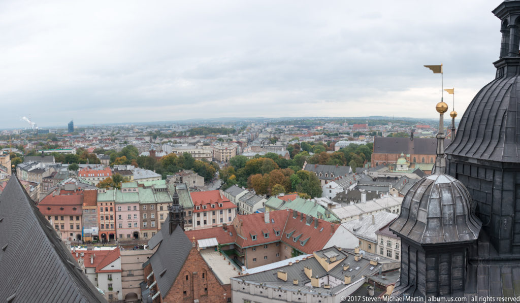 Elevated Krakow panorama framed by dark basilica roofs and a small cupola with gilded weather vane above dense historic rooftops.