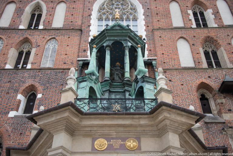 Green-patinated copper canopy with columns and sculpture projecting from the red-brick façade beneath Gothic windows.