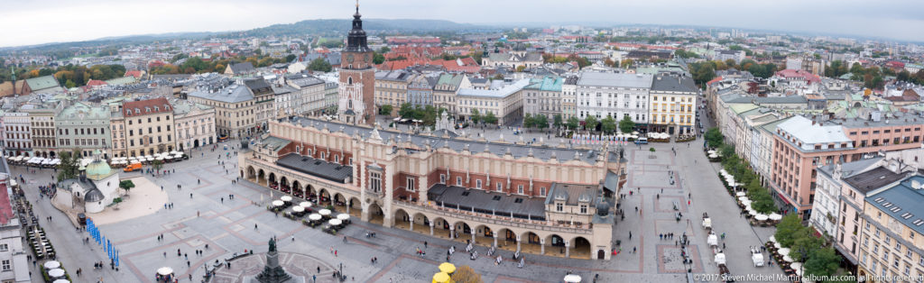Wide elevated panorama of Krakow Main Market Square with Cloth Hall, Town Hall Tower, surrounding townhouses, and pedestrians below.