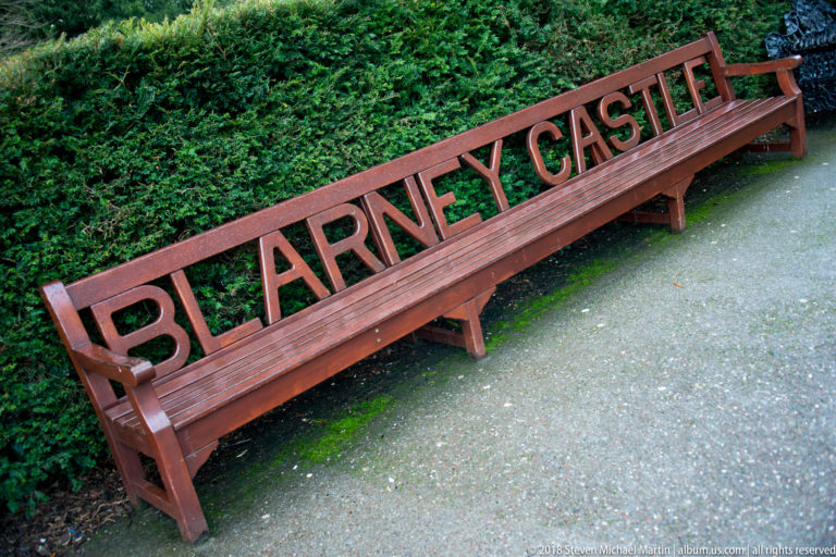 Long wooden bench with the words BLARNEY CASTLE cut into the backrest, set beside a dense green hedge.