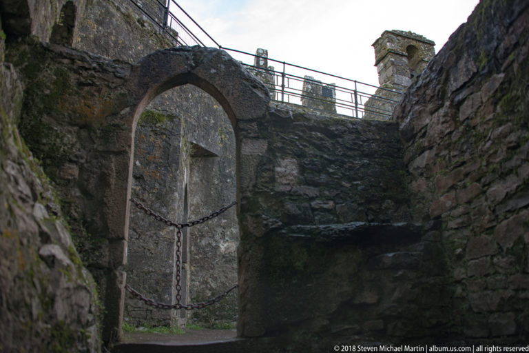 Dark stone passage with tall arched doorway, heavy chain barrier, rough walls, and modern railings above.