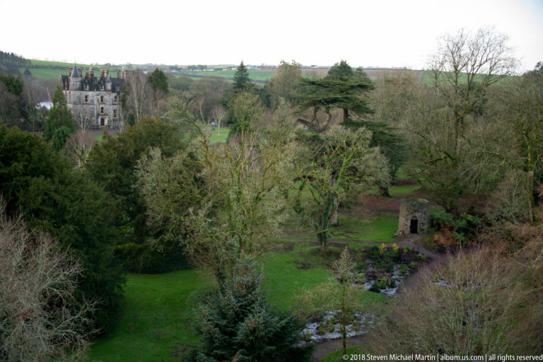 Elevated view over Blarney Castle gardens with lawns, mature trees, a small stone structure, and a distant castellated mansion.