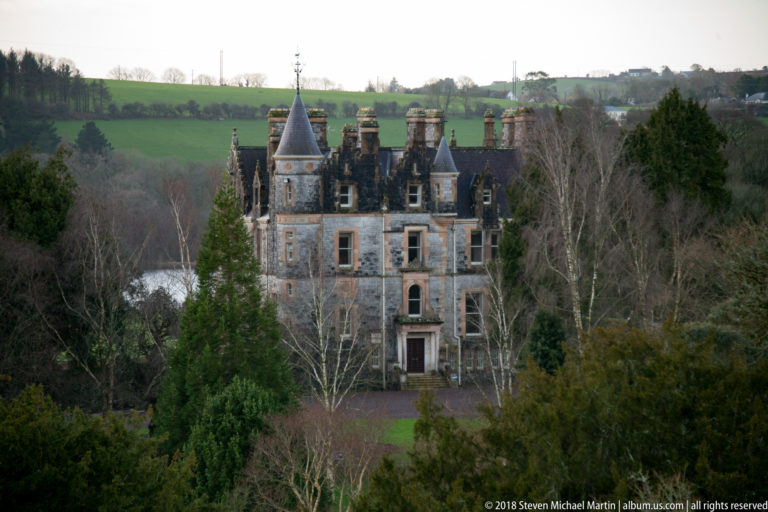 Distant castellated mansion with turrets, steep roofs, chimneys, and central entrance, partly framed by trees.
