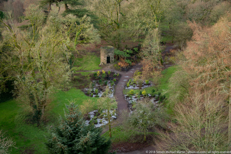 High-angle view of a curving garden path, dense plantings, and a small stone structure on the Blarney Castle grounds.