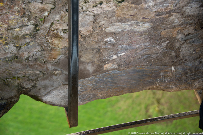 Close view of rough weathered stone at an opening, crossed by dark metal safety rails with green lawn visible below.